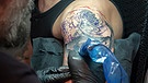 Ein Tätowierer bringt am Oberarm eines jungen Mannes ein buntes Tattoo auf. | Bild: picture-alliance/dpa