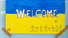 RCHIV - 09.03.2022, Bayern, München: Ein Schild in den Farben der Ukraine mit der Aufschrift "Welcome" ist am Hauptbahnhof zu sehen. Im Hintergrund werden Flüchtlinge aus der Ukraine nach ihrer Ankunft von Mitarbeitern der Caritas und freiwilligen Helfern empfangen. | Bild: Sven Hoppe/dpa +++ dpa-Bildfunk +++