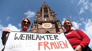 Zwei Frauen der Initiative Maria 2.0 der Katholische Frauengemeinschaft in der Erzdiözese Freiburg halten vor dem Münster ein Banner mit der Aufschrift "Weiheämter auch für Frauen" in die Höhe.  | Bild: dpa-Bildfunk/Patrick Seeger