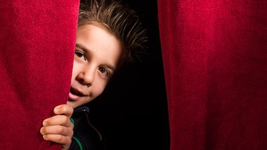 Ein Kind schaut zwischen einem roten Vorhang hervor. | Bild: stock.adobe.com/Deyan Georgiev