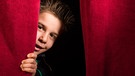 Ein Kind schaut zwischen einem roten Vorhang hervor. | Bild: stock.adobe.com/Deyan Georgiev