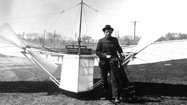 Mann in Kleidung des 19. Jahrhunderts steht vor dem ersten Motorflugzeug 1901 | Bild: picture-alliance/dpa