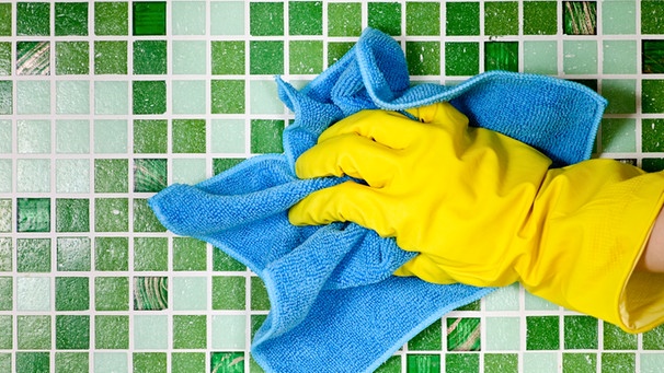 Eine Hand im gelben Gummihandschuh wischt mit blauem Tuch über grüne Mosaikfliesen | Bild: colourbox.com