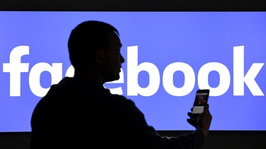 Ein Mann steht mit seinem Smartphone vor einem Facebook-Logo auf einem Monitor.  | Bild: dpa-Bildfunk/Carsten Rehder