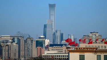 China, Peking: Rote Fahnen flattern auf der Großen Halle des Volkes, während im Hintergrund Bürohochhäuser des Central Business District zu sehen sind. | Bild: dpa-Bildfunk