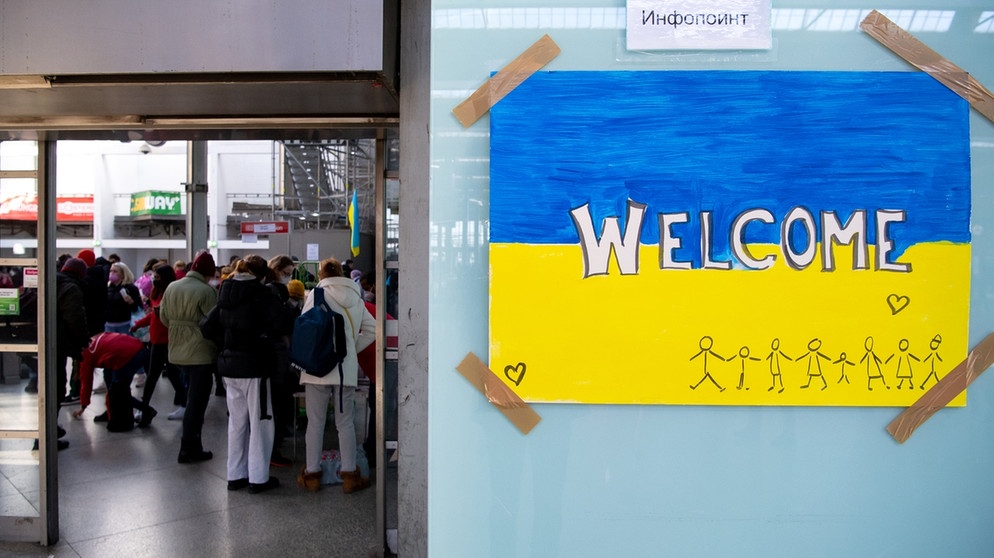 Ein Schild in den Farben der Ukraine mit der Aufschrift "Welcome" am Münchner Hauptbahnhof. Im Hintergrund werden Geflüchtete nach ihrer Ankunft von Mitarbeitern der Caritas und freiwilligen Helfern empfangen | Bild: dpa-Bildfunk/Sven Hoppe