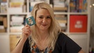 Die Spieleentwicklerin Rita Modl hält lächelnd eine Spielkarte hoch. | Bild: BR/Andreas Krieger