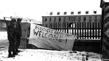Zwei Soldaten betrachten im KZ Flossenbürg einen Banner mit der Aufschrift "PRISONERS HAPPY END! WELCOME!" | Bild: National Archives, Washington DC (NARA)