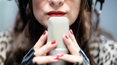 Rote Frauenlippen vor einem Mikrophon, das die Frau mit ihren Händen und roten Nägeln umgreift | Bild: picture alliance/Matthias Balk/dpa