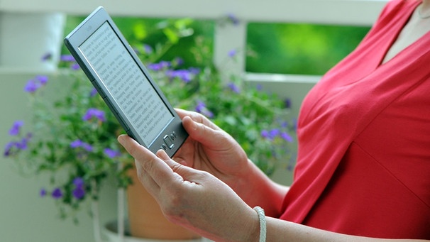 Eine Frau sitzt auf ihrem Balkon und liest mit ihrem E-Book-Reader in einem E-Book. | Bild: picture-alliance/dpa
