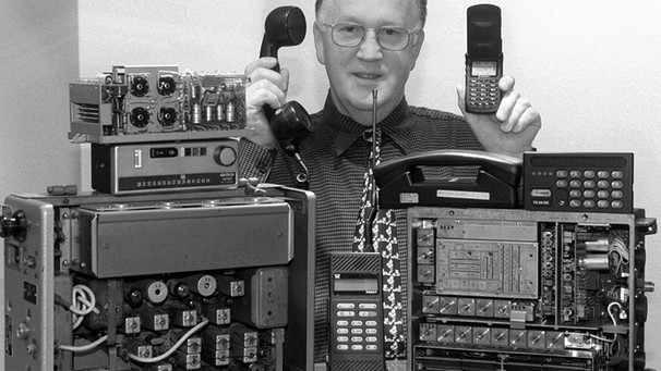 Telekom-Mitarbeiter Heinz Sänger hält in seiner rechten Hand den Hörer eines rund 30 kg schweren A-Netz-Telefons und ein rund 125 Gramm leichtes D-Netz-Handy in seiner linken.  | Bild: picture-alliance/dpa