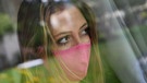 Eine junge Frau mit pinker Schutzmaske steht am Fenster in einem Zimmer und blickt wehmuetig, traurig in die Ferne. | Bild: picture alliance / Sven Simon