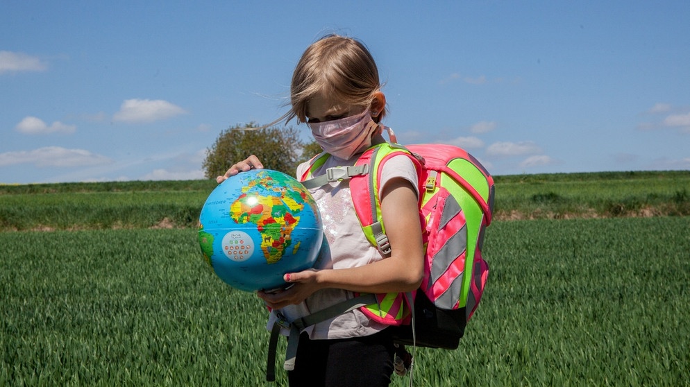 Kind mit Nasen-Mund-Schutz gegen das Coronavirus auf dem Schulweg mit einer Weltkugel in der Hand: Sind Kinder weniger von SARS-CoV-2 betroffen? | Bild: picture alliance/Fotostand