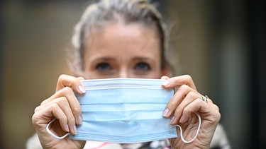 Eine Frau setzt einen Mund-Nasen-Schutz auf. | Bild: picture-alliance/dpa