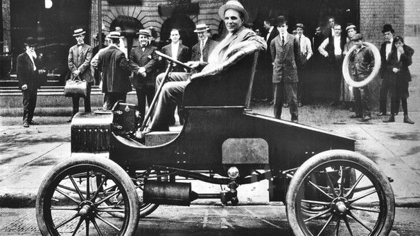 Der 37jährige Henry Ford posiert im Jahr 1900 vor seinem Werk in Detroit (Michigan) am Lenkrad eines brandneuen Autos | Bild: picture-alliance / dpa