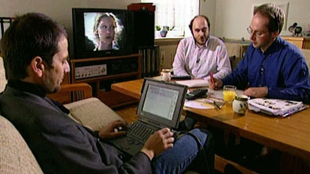 Drei Personen (v.l.n.r.: Wolfgang Hartmann, Elmar Dosch, Bernd Benecke) sitzen um einen Tisch, arbeitend, einer mit einem Laptop, andere nachdenkend und schreibend. Im Hintergrund ein Fernsehbildschirm | Bild: Screenshot