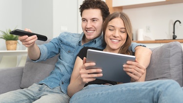 Mann mit Fernbedienung in der Hand, Frau mit Tablet in den Händen sitzen auf einer Couch | Bild: colourbox.com