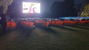 viele rote Liegestühle bei Regen, draußen auf dem Olympiagelände, vor großer Kinoleinwand. Zuschauer sitzen vorwiegend mit geöffnetem Regenschirm auf den Stühlen | Bild: Beate Hornung