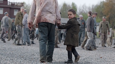 Ein Kind geht im KZ-Gelände an der Hand eines Mannes und schaut sich um. | Bild: BR/MDR/UFA Fiction