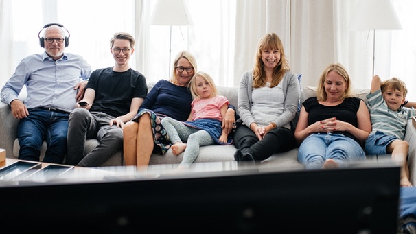 Eine Familie sitzt auf dem Sofa und schaut gemeinsam Fernsehen. Ein Familienmitglied hört die Originalsprache zur laufenden Sendung über Kopfhörer. | Bild: BR/Julia Müller