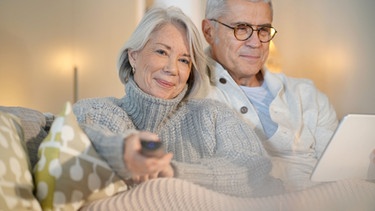 Ein älteres Ehepaar sitzt auf der Couch. Die Frau sieht fern, der Mann blickt auf sein Tablet. | Bild: stock.adobe.com/goodluz