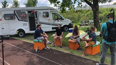 Das PULS-Team dreht für die Festivalheimat im Corona-Sommer 2020 vor dem PULS Wohnmobil in Perlesreut. Vier junge Leute sitzen auf improvisierten Kisten vor dem Wohnmobil und werden von einem Kameramann gefilmt. | Bild: Emanuel Abréu