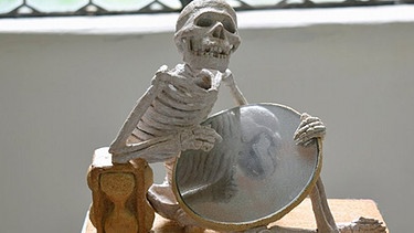 Kleine sitzende Skelett-Figur hält einen runden Spiegel  / Ausstellung "Totentanz" in der Kartause Buxheim | Bild: Manfred Mühlbauer