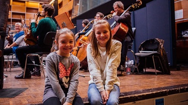 Zwei Mädchen sitzen auf Bühnenrand vor Orchester | Bild: BR / Max Hofstetter