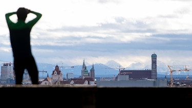 Person blickt auf Skyline von München mit Baukränen vor Alpenpanorama. | Bild: dpa-Bildfunk/Sven Hoppe